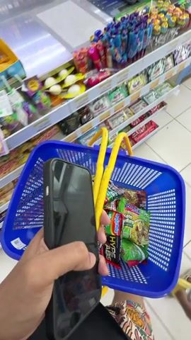 The video opens with a close-up of a person's hand holding a smartphone. The phone screen is dark. In the background, a blue shopping basket is visible, filled with several packets of instant noodles. Two bright yellow plastic handles of the basket extend upwards. Behind the basket, shelves are stocked with various colorful products, including what appear to be small bottles and packets. A payment terminal or scanner is partially visible on a counter to the left. The overall environment suggests a retail setting, possibly a convenience store or small supermarket, with a focus on food items.