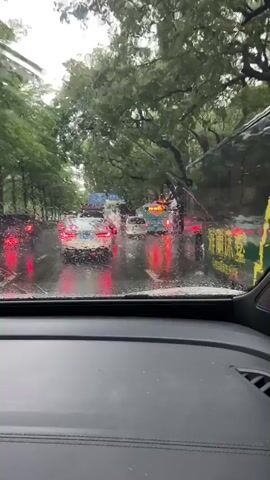 The video shows a view from inside a vehicle looking through a rain-streaked windshield at a traffic scene. It is raining heavily, with water droplets visible on the glass. Several cars and a bus are on a wet, tree-lined road. The vehicles' taillights are illuminated, reflecting on the wet asphalt. The traffic appears to be moving slowly or stopped. The overall environment is overcast and appears to be daytime.