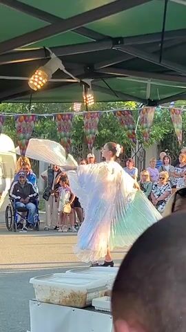 A female dancer in a white iridescent costume with flowing sleeves and a pleated skirt performs on a raised platform under a green tent. She is facing away from the camera, with her arms extended. Several strings of colorful balloon-shaped decorations hang overhead, along with spotlights. A crowd of people, including a man in a wheelchair, watches the performance from behind a low barrier. In the foreground, a table with plastic food containers is visible. The scene appears to be an outdoor event.