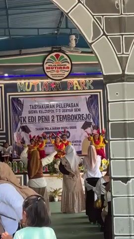A group of young children in red and yellow costumes are performing a dance on a stage. The stage is decorated with banners that read "MUTIARA PARK" and "TASYAKURAN PELEPASAN SISWA KELOMPOK G GEBYAR SENI TK EDI PENI 2 TEMBOKREJO TAHUN PELAJARAN 2025/2026". Several adults are watching the performance from the audience, some of whom are wearing hijabs. The environment appears to be outdoors, possibly at a park or school event. The lighting is bright, suggesting it is daytime. The overall atmosphere seems festive and celebratory.