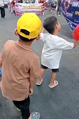 Two young boys are walking in a street with a parade in the background. The boy in the foreground wears a tan t-shirt and black pants, with a yellow baseball cap on his head. He is looking away from the camera, his arms slightly raised. The boy behind him wears a white t-shirt and dark shorts, with sandals on his feet. He is holding a red and white inflatable object, possibly a beach ball, and looking up and to his right. In the background, a banner reads "REPUBLIK INDONESIA". Another vehicle with text including "SMK PELAYARAN OPERASI LAUT" is visible. The street appears to be paved, and there are other people and decorations associated with a parade. The lighting suggests it is daytime.