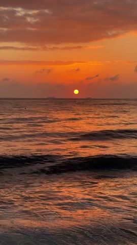 A video captures a sunset over the ocean. The sun, a bright orange orb, is visible just above the horizon, casting a warm glow across the sky and water. The sky is filled with wispy clouds, tinged with shades of orange and pink. Gentle waves roll towards the shore, their surfaces reflecting the colors of the sunset. In the distance, faint silhouettes of landmasses or ships can be seen. The overall mood is serene and tranquil.