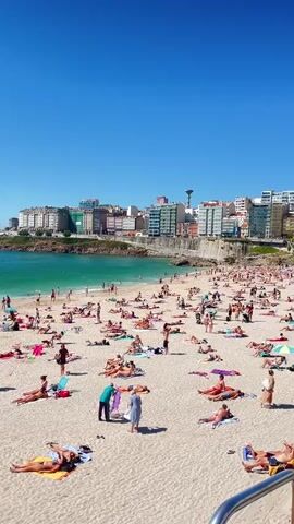 The video shows a crowded beach on a sunny day. Numerous people are spread out on the sand, many sunbathing on towels or reclining on chairs. Some individuals are walking or standing, while others are seen in the shallow water near the shore. In the background, buildings line the coast, and the turquoise water stretches out to the horizon under a clear blue sky. There are trash cans visible on the beach.