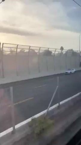 A video shot from a moving vehicle shows a highway with several cars traveling on it. The highway is bordered by a tall grey fence. Beyond the fence are trees and bushes, and the sky is visible with a mix of blue sky and clouds. The light suggests it might be morning or evening. The perspective suggests the camera is looking down at the highway from an elevated position, possibly from another road or a train.