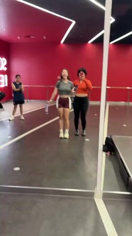 The video shows a dance studio with red walls and mirrors. In the foreground, two young women are posing for a selfie in a large mirror. The woman on the left is wearing a gray crop top and maroon shorts. The woman on the right is wearing an orange long-sleeved crop top and black leggings. In the background, another young woman is standing in a dark blue t-shirt and black shorts, facing away from the camera. There are railings along the wall behind her. The floor is dark gray with white lines.