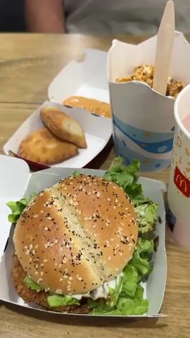 A medium shot captures a wooden table laden with fast food from McDonald's. In the foreground, a crispy chicken sandwich sits in its white paper box, topped with sesame seeds and lettuce. To its right, a pink beverage in a McDonald's cup is partially visible. Behind the sandwich, two empanadas lie in a red-and-white cardboard container, accompanied by a sauce packet. Further back, a white cup with a blue wave pattern holds popcorn and a wooden utensil. The background is blurred, showing a person in a light-colored t-shirt and a black strap across their chest. The lighting is bright and even, suggesting an indoor setting.