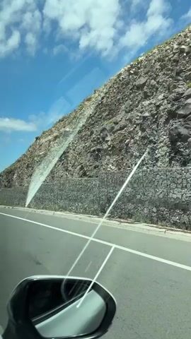 This is a first-person perspective video taken from inside a vehicle, likely a car, driving along a paved road. The road has a white dividing line. In the foreground, a side-view mirror of the car is visible.

The primary subject is the scenery outside the car. To the right of the road, a steep, rocky hillside rises sharply. The hillside is covered with rocks of various sizes and some sparse vegetation. A gabion wall, constructed from wire cages filled with stones, is built along the base of the hillside, likely to prevent erosion or rockfalls. Sunlight is reflecting off the windshield, creating diagonal lines across the frame.

The sky is a bright blue with scattered white clouds. The overall impression is a sunny day. The landscape appears natural and somewhat rugged, suggesting a mountainous or hilly region. The presence of the road and the gabion wall indicates human infrastructure interacting with the natural environment.

There is no visible text within the video. The activity is simply driving along a road with a view of the natural landscape. The time of day appears to be daytime, given the bright sunlight. The weather seems to be clear and sunny. The location context is Mastara, Armenia. The video conveys a sense of travel and observation of the surroundings.