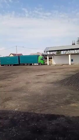 A bright green semi-truck with two teal-covered trailers is parked in a lot. To the left of the truck, a yellow front-end loader is moving across the scene. The ground is covered in dark dirt or gravel. In the background, there are buildings and trees. The sky is blue with some clouds.