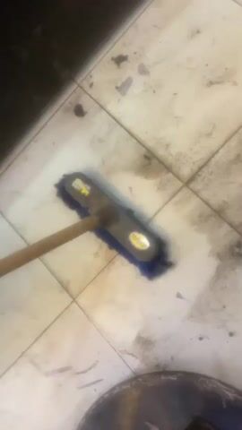 A handheld video shows a blue-bristled broom with a wooden handle being used to sweep a tiled floor. The floor appears dirty with dark smudges and streaks. The broom is in motion, sweeping across the tiles. The lighting is dim. The action is focused on the sweeping of the floor.