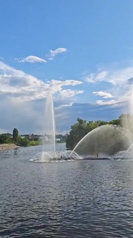 This video showcases a large fountain display in a body of water, likely a river or lake, under a bright blue sky with scattered white clouds. The sun is high, suggesting it is daytime, possibly late afternoon given the warm light. The fountain jets are powerful, creating impressive arcs and sprays of water that catch the sunlight. In the background, a lush green treeline borders the water, and hints of buildings are visible on the distant shore. The overall scene evokes a sense of tranquility and natural beauty, enhanced by the dynamic play of water and light. No text is visible in the video.