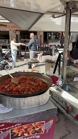 The video shows an outdoor market in a European town. The first shot features a food stall with a man standing behind it, serving food from a large pan and a display case. A second man stands nearby, apparently making a purchase. The camera then pans to follow people walking through the market. Some are pushing strollers, others are window-shopping. The market has many stalls, some selling sausage, others selling terrines. The market appears to be crowded, on a sunny day.  The buildings in the background are painted in pastel colors.