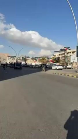 The video shows a black van driving down a road with a police motorcycle escort. The van has flashing blue and red lights on its roof. The road is lined with parked cars and buildings. There are people on motorcycles riding alongside the van. The sky is blue with white clouds. There is a person standing on the sidewalk to the right of the road, partially visible at the bottom of the frame.