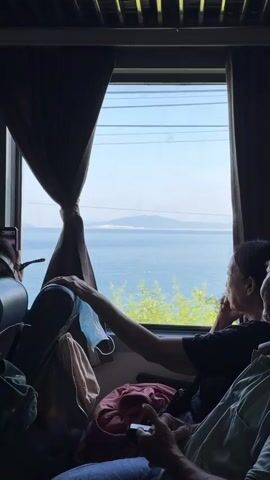 This media file captures a view from inside a train traveling through Dai Lanh, Vietnam. The scene is framed by dark brown curtains on either side of a large window. Outside, the bright daylight reveals a coastal landscape. The sea, a deep blue, stretches to the horizon where a hazy, light-colored landmass, likely an island or distant coast, is visible under a clear, pale blue sky. Lush green trees and foliage partially obscure the view of the water, indicating the train is passing close to the shore.

Inside the train, several people are seated. In the foreground and mid-ground, passengers are visible from behind or the side, mostly looking out the window or attending to their devices. One woman in the mid-ground, with her arm resting on the seatback, gazes intently at the passing scenery. A light blue surgical mask is hanging from her arm. Another passenger in the foreground is holding a phone, possibly also looking at it or taking a picture. Further back, another person is wearing large headphones. Luggage, including a pink bag and a green backpack, is visible on the seats and floor, suggesting a journey in progress.

The time of day appears to be late morning or early afternoon, given the bright sunlight and clear sky. The weather seems pleasant, suitable for travel and enjoying the coastal views. The presence of the sea, the greenery, and the style of the train interior are indicative of the Dai Lanh region of Vietnam. The overall mood seems to be one of quiet observation and travel, with passengers engrossed in their individual experiences of the journey and the scenery. No visible text is present in the media.