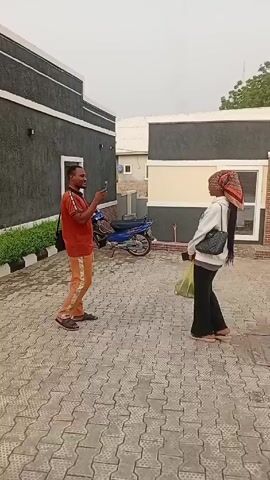 A man and a woman are standing facing each other in an outdoor paved area with buildings in the background. The man is wearing an orange t-shirt, tan pants with a side stripe, and slides. He is holding a smartphone and appears to be taking pictures of the woman. He is smiling. The woman is wearing a white hooded sweatshirt, black pants, and slides. She also has a patterned headscarf and is holding a yellow shopping bag. A blue motorbike is parked in the background. The buildings are painted grey and tan, and there is some greenery along the wall of one of the buildings. The scene is well-lit, suggesting daylight.
