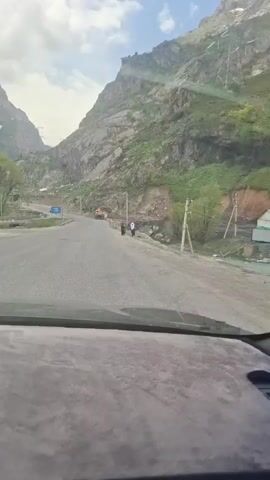 The view from inside a car shows a road winding through a mountainous landscape. The mountains are rocky and partially covered with green vegetation. An orange bulldozer is parked on the side of the road. Further ahead, three people dressed in traditional clothing are walking near the road. Power lines and utility poles are visible along the road and on the mountainside. The sky is partly cloudy. The road appears to be mostly clear, though it has some gravel on the surface. A barrier lines the side of the road next to a creek bed.