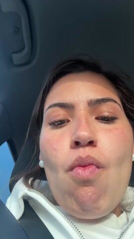 A woman is sitting in the driver's seat of a car, wearing a white hooded sweatshirt and pearl earrings. The seatbelt is visible. She is making faces for the camera, sticking out her tongue, smiling, and puckering her lips. The car's interior is visible, with the headliner and window frame in view. Light from outside the car illuminates the woman's face. Music is audible in the background.