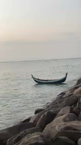 The video shows a boat floating on the water near a rocky shore. The boat is dark, with a light blue stripe along the top. It has pointed ends that curve slightly upwards. The water is calm, with a few other objects visible in the distance. The sky is overcast, and the light suggests it could be early morning or late afternoon. The rocks on the shore are large and dark. There are no people visible in the video.