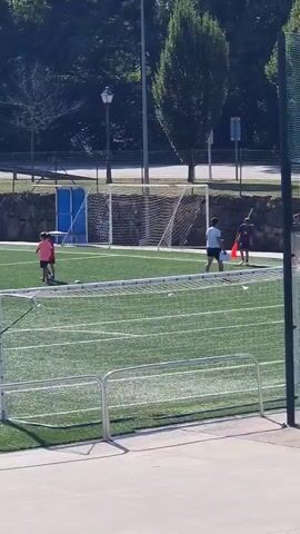 The video shows a soccer field on a sunny day. There are several young people on the field. Some are wearing pink shirts, while others are in darker colored shirts. There are two soccer goals visible, with nets. There are also some white balls scattered on the field, some of which are inside the goals. One person in a dark shirt appears to be kicking a ball. The field is green artificial turf, and there are trees and a stone wall in the background. There is also a fence in the foreground.