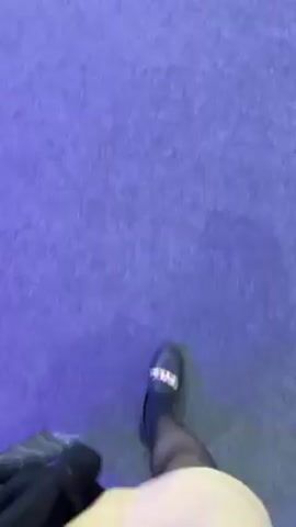 The video begins with a view of a person’s feet as they walk on a purple carpet. The person is wearing black tights and black Crocs with a silver chain. The person is carrying a black bag. The camera then pans up to show a crowded exhibition hall. Many people are walking around on a grey carpet. There is a sign on the carpet that reads “F”. The camera then focuses on a white car that is on display behind a glass barrier. The car has orange rims. The word “APTIV” is written on the side of the car. People are standing around the car, looking at it and taking pictures.