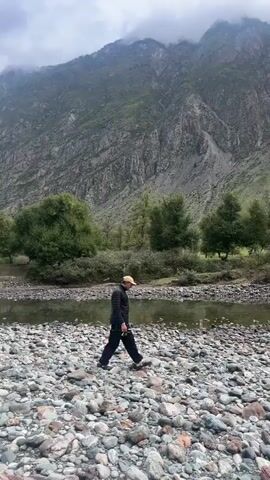 A man walks along a rocky riverbank with a mountain in the background. The man is wearing a black jacket, dark pants, and a tan hat. He is walking from left to right across the frame. The river is shallow and clear. The mountain is steep and covered in vegetation. The sky is cloudy.