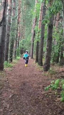 A young child, wearing a gray hooded jacket, blue jeans, and bright yellow rain boots, runs towards the camera on a dirt path in a forest. The child is holding a clear umbrella with a blue pattern open, twirling it as they run. Their face is visible, and they appear to be smiling or laughing. The path is lined with tall, slender pine trees, and the ground is covered in pine needles and fallen leaves. The scene suggests a damp or rainy day, fitting with the child's attire and the presence of the umbrella. The overall mood is playful and joyful, as the child seems to be enjoying their walk or run through the woods. The location is identified as Русский Вожой, Russia.