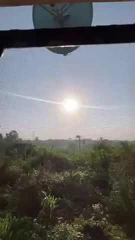 A hand reaches towards a bright sun seen through a window. The window has a screen and iron bars. Outside the window, greenery is visible, along with a satellite dish mounted above.