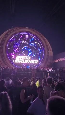The video captures a nighttime outdoor concert or festival. A large circular LED screen dominates the background, displaying abstract purple and blue patterns with the name "BRAD BRUNNER" in illuminated text. Blue spotlights emanate from the top of the screen, casting beams into the darkness. A crowd of people is gathered in front of the stage, silhouetted against the bright lights. Some individuals are standing, while others appear to be sitting or leaning. The atmosphere suggests a lively event.