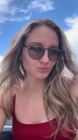 A woman with long blonde hair, wearing sunglasses and a red tank top, poses for a selfie. She smiles and runs her hands through her hair. Behind her is a blue sky with scattered clouds and a boat.