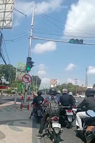 A group of motorcyclists are stopped at a traffic light on a sunny day. The camera is mounted on a motorcycle, looking forward. The traffic light is green. The motorcyclists are wearing helmets and casual clothing. Some of the motorcycles have backpacks. The road is paved and there are white lines indicating lanes and a pedestrian crossing. There are buildings and trees in the background. The scene appears to be in a city or town.