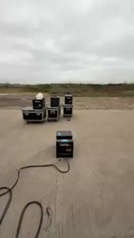 The video shows a black box-shaped machine sitting on an open concrete surface with a long black cable connected to it. The machine shoots out a large burst of flames. Behind the machine are several black storage trunks of varying sizes. In the background, there is a flat landscape with sparse vegetation and a cloudy sky.