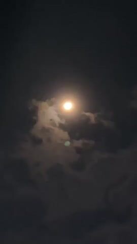 A nighttime scene with a bright full moon centered in the frame. The moon is partially obscured by dark, wispy clouds that are illuminated by the moonlight. The sky is dark. A slight green lens flare is visible near the bottom center of the frame. The bottom edge of the frame shows a hint of a white structure, possibly a building.