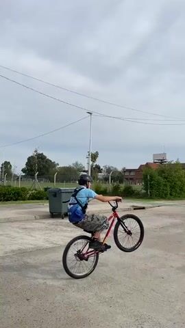 A person wearing a helmet, a blue shirt, and camouflage shorts is riding a red bicycle on one wheel, doing a wheelie. The cyclist is on a concrete surface with street lamps and overhead wires. There are green trash bins nearby. In the background, there is a low brick building surrounded by trees and a tall communications tower. The sky is overcast. The cyclist continues down the street, maintaining the wheelie.