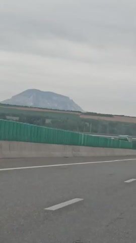The video shows a view from the perspective of someone in a moving vehicle on a highway. The road is paved with white lane markings and guardrails are visible along the side. In the distance, there are hills and fields, with some trees and buildings scattered throughout the landscape. The sky is overcast. A blue sign is visible on an overpass in the first frame.