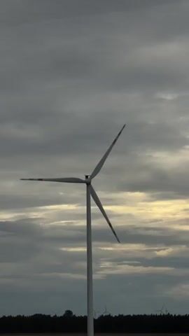 The video shows a single wind turbine against a cloudy sky. The turbine is tall and white with three blades. The blades are spinning, indicating that the wind is blowing. The sky is overcast with dark gray clouds, but there are some lighter patches of sky visible, suggesting that the sun might be behind the clouds. In the background, there is a dark line of trees and some other wind turbines visible in the distance. The overall scene is one of a windy, perhaps stormy, day.