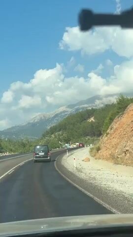 A black van drives down a paved road. The road is curved and has a white dividing line. On the right side of the road is a rocky hillside with green trees. In the background are mountains covered in trees and a blue sky with white clouds. There are other vehicles on the road.