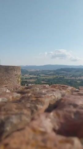 The media file captures a scenic view from an elevated vantage point, likely a historical structure given the stone wall in the foreground. The scene looks out over a Tuscan landscape characterized by rolling hills, lush greenery, and scattered buildings with red-tiled roofs, indicative of a European village or small town. In the middle ground, there's a cluster of houses and trees, suggesting a settlement nestled within the countryside. The distant background features more undulating hills and a hazy horizon, under a clear blue sky with a few wispy white clouds.

The time of day appears to be late morning or early afternoon, with bright sunlight casting shadows and illuminating the landscape. The weather seems pleasant and clear, suitable for outdoor sightseeing. The foreground is dominated by a rough, reddish-brown stone surface, possibly a wall or walkway, with a cylindrical stone structure on the left, resembling part of a tower or ancient fortification. To the right, a row of green trash receptacles is visible along a paved pathway, adding a modern element to the otherwise historical and natural setting.

The overall mood evoked is one of tranquility and picturesque beauty, typical of the Italian countryside. There is no visible text in the media file. The setting is identified as Peccioli, Italy, which aligns with the visual characteristics of the Tuscan region. No people are visible in the frame. The activity, if any, is simply the presentation of a static vista.