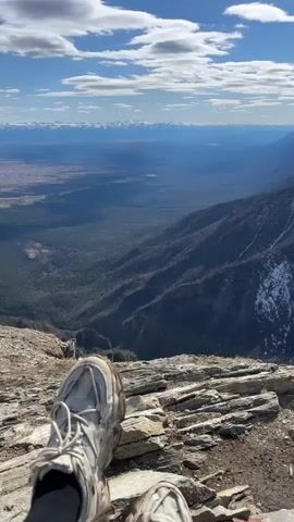 The video shows a scenic mountain view, seen from a high vantage point, likely a mountain peak. The foreground is composed of rocky terrain, with a pair of light-colored hiking shoes visible, suggesting someone is sitting and enjoying the view. The middle ground showcases a valley with dense forestation, while the background reveals a series of mountain ranges, some with visible snow cover. The sky is bright blue with scattered white clouds, and the sun is shining. The camera pans up, emphasizing the vastness of the landscape and the height of the viewing point.