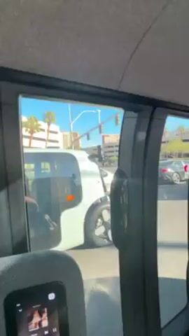 The video, shot from inside a vehicle, reveals a partial view of its interior. The camera angle is low, capturing the side window, a portion of the seat, and the sunroof. The interior is a muted grey, with a clean, modern design.  Through the window, one can see a light-colored vehicle with a distinctive design waiting at a traffic light. The scene outside features a city street with palm trees, buildings, and a crosswalk. A Nordstrom store sign is visible in the distance. The sky is clear and blue, and the overall lighting suggests a bright, sunny day. Music is playing in the background.