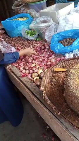 A person wearing a blue hijab and a dark blue abaya is standing behind a market stall. Their hands are visible, and they are sorting through a large pile of red onions. The onions are spread across the wooden surface of the stall. To the left of the onions, there are several blue plastic bags filled with various goods, including what appears to be dried spices or grains. Behind the onions, there are more plastic bags, some containing green leafy vegetables and others containing yellow items that could be dried fruit or chips. To the right of the onions, a woven basket is partially visible, filled with a brown, granular substance. The stall appears to be outdoors or in an open-air market, with a rough wooden structure and a concrete floor visible.