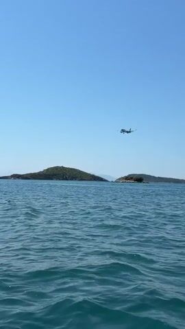 A passenger airplane is in mid-flight over a body of water. The plane is white with dark markings and appears to be descending, likely for landing. The water is a vibrant blue-green, with gentle ripples on its surface. Two small, tree-covered islands are visible in the background, positioned on either side of the plane. The sky is clear and a bright blue, indicating fair weather. The overall scene suggests a scenic coastal or island location under a sunny sky.