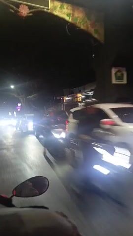 The video shows a night-time view from the perspective of someone riding a motorcycle on a street. Several cars are visible, some with their headlights on, creating a blurred effect due to motion. The environment appears to be urban with buildings and trees lining the street. The overall impression is of a busy road at night with moving vehicles.