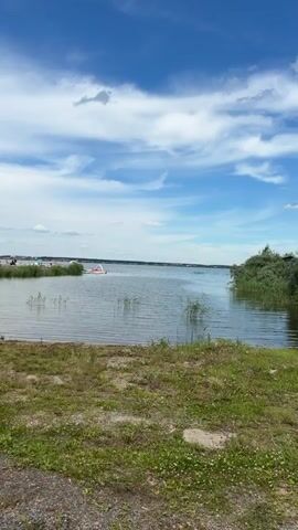 The video shows a body of water with reeds growing along the shore. In the foreground, there is a grassy area with a few boats, some of which are green pedal boats. In the background, there are buildings and a few people can be seen on the shore and in the water. The sky is blue with white clouds. The overall scene appears to be a park or recreational area by a lake or river on a sunny day.
