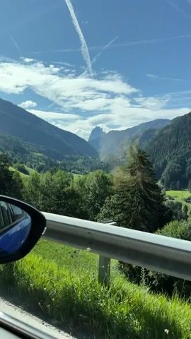 The video shows a view from inside a moving vehicle, looking out the side window. A car's side mirror is visible on the left side of the frame, reflecting the sky and clouds. The landscape outside is mountainous and covered with lush green trees. A metal guardrail runs along the edge of the road in the foreground. In the distance, a valley is visible with more trees and a sharp mountain peak piercing the sky. The sky is a clear blue with scattered white clouds, some of which are crossed by faint white lines, possibly contrails from airplanes. The overall scene depicts a scenic drive through a mountainous region.