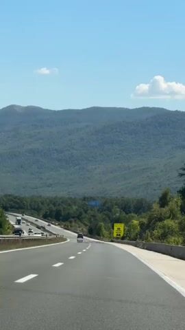 The video shows a highway with cars and trucks traveling on a sunny day. The highway curves to the right and is surrounded by green trees and mountains in the background. A yellow sign with a picture of cars on a bumpy road is visible on the right side of the highway. The sky is blue with a few white clouds.