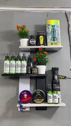 A video pans across a wall-mounted shelving unit in what appears to be a barber shop or grooming station. The shelves hold an assortment of hair products, styling tools, and decorative plants. On the top shelf, there are two small artificial plants in white pots, a black bottle, a yellow and black electric trimmer, and a box with an image of a man and a green electric clipper. The middle shelf displays several identical green spray bottles labeled "HAIR TONIC", a few smaller dark bottles, and another artificial plant in a white pot. To the right of the middle shelf, a black spray bottle and a yellow-handled tool are visible. The bottom shelf features a blue and red tin labeled "STYLING CLAY", a black tin with "HAIR POMADE" printed on it, several more green spray bottles of "HAIR TONIC", and a small clear bottle. The background is a grey, textured wall.