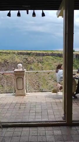 The video shows an outdoor restaurant with a scenic view of a canyon. A waiter, wearing a light blue shirt and black pants, is serving a table with two women. One woman, wearing a light blue top and tan pants, is seated on the left, facing away from the camera. The other woman, wearing a pink and white striped shirt, is seated on the right, facing the table. The table has food and drinks on it. The restaurant has a tiled floor and a decorative railing overlooking the canyon. The sky is cloudy.
