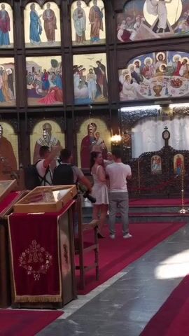 The video shows the interior of a church. In the foreground on the left, a red cloth covers a wooden stand with a glass container on top. A wooden chair is positioned near the stand. In the center of the frame, two men and a woman are interacting. One man, wearing a white t-shirt and grey pants, holds a baby. The woman, in a white dress, is embracing the man holding the baby. Another man, with a backpack, stands to their left, facing them. Behind them, the church walls are adorned with religious icons and paintings. The floor is covered with a red carpet. The lighting appears to be natural, coming from the windows of the church. The overall atmosphere seems to be a family gathering or a religious ceremony.