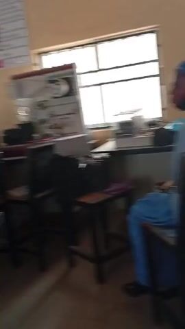 The video depicts a classroom scene with several individuals seated at desks. Most are facing towards the front of the room, where a projection screen is visible. The people appear to be of diverse ethnicities and are dressed in a variety of clothing styles, including traditional attire. Some individuals have laptops open on their desks, suggesting they may be engaged in a lesson or presentation. The classroom has a somewhat cluttered appearance, with various objects scattered around, including bags and water bottles. The room is lit by ceiling lights and natural light coming through the windows.