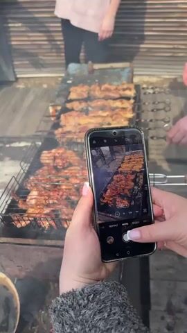 A person is taking a picture of meat cooking on a grill with their smartphone. The grill is full of skewers and a grate covered with meat. Smoke is rising from the grill. Other people are visible in the background. The phone screen shows the live view of the meat cooking on the grill. The phone is being held steadily with both hands. The person is wearing a gray sweater and has manicured nails.