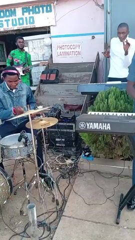 The video shows an outdoor performance with a live band and people dancing. The band consists of a drummer wearing headphones, a keyboard player next to a Yamaha keyboard, and a guitar player in a green shirt.  There are speakers and equipment set up around them. A sign for "FREEDOM PHOTO STUDIO QUALITY GUARANTEED" and another for "PHOTOCOPY LAMINATION" are visible in the background. The environment appears to be a street with people passing by.  Several people are dancing to the music. One person wearing sunglasses, a white jacket, and jeans is moving in a rhythmic way.